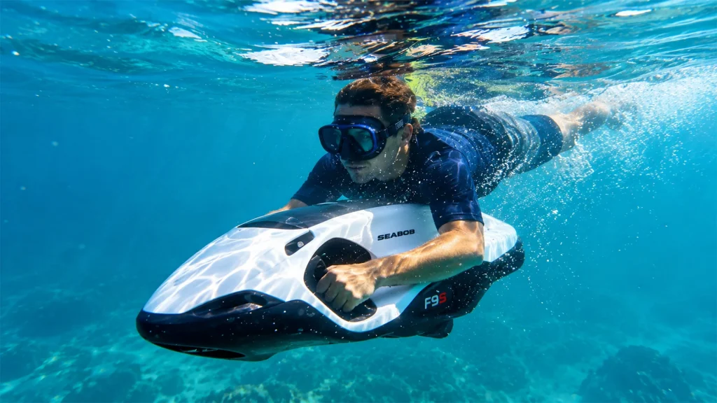 Man wearing a diving mask riding a white SEABOB F9 S underwater scooter through clear turquoise water.
